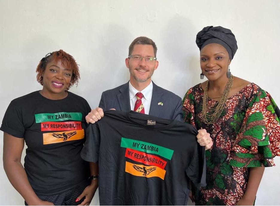 Two women and a man pose for a picture with a My Zambia, My Responsibility shirt displayed