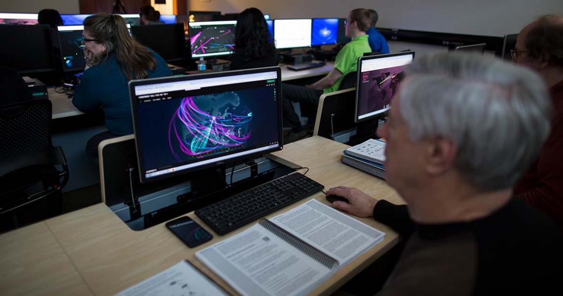 An older white man sits at a computer in a classroom.