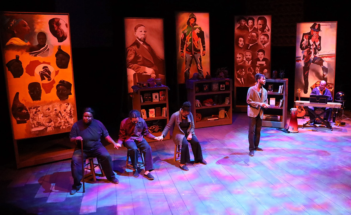 Three people seated on a purple stage with a fourth person at a microphone, in front of orange painted scrim panels.