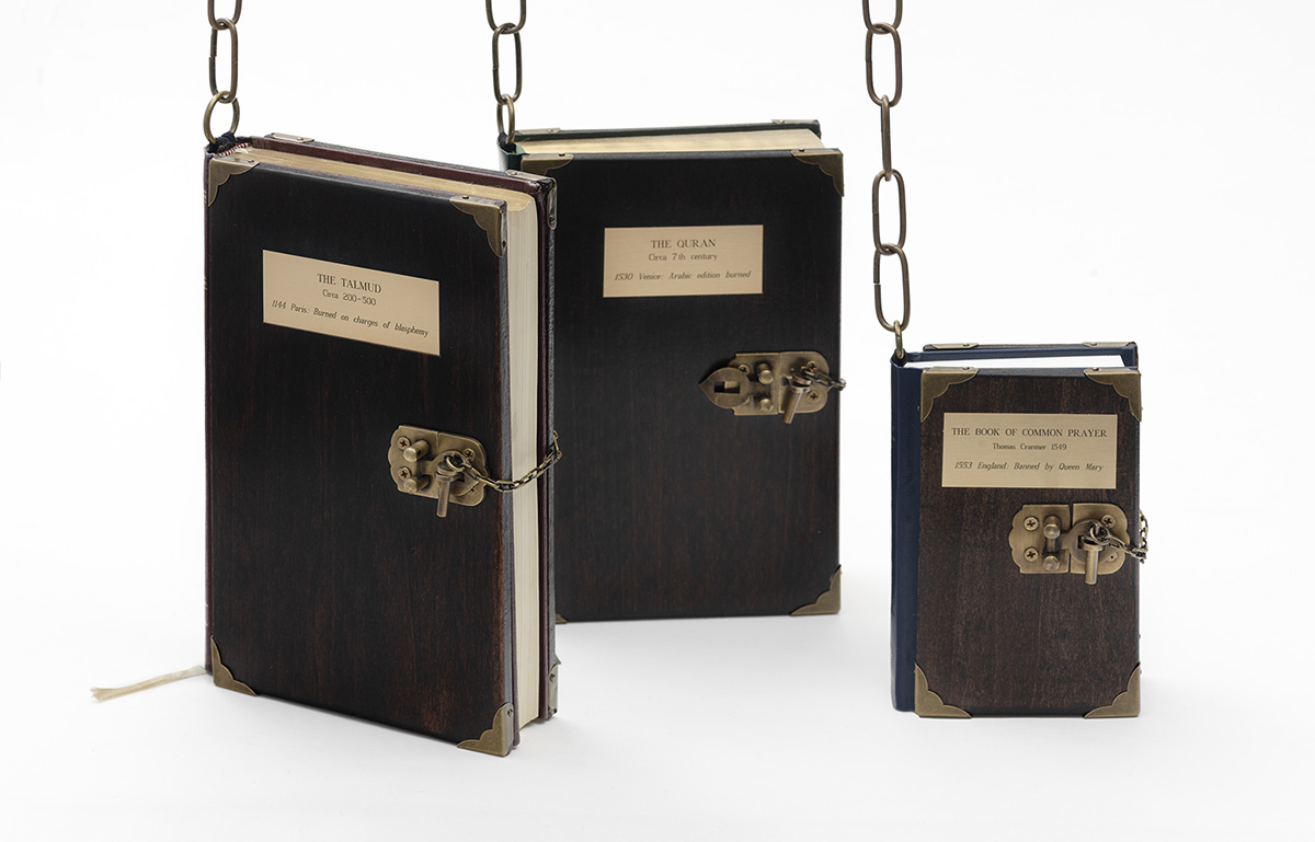 A series of leather bound books with clasped locks dangling from chains: The Talmud, The Quran, and The Book of Common Prayer