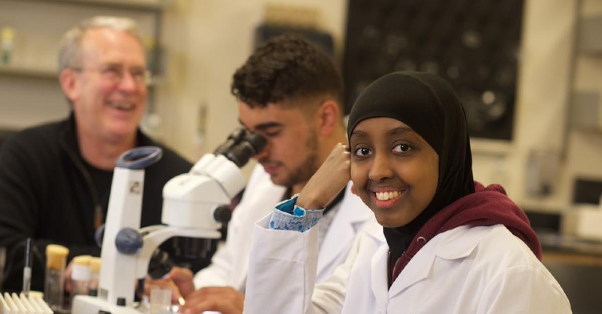 Two students in white lab coats in lab with microscope and faculty