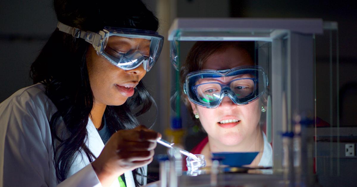 An instructor looks on as a student adds liquid to a bottle in a lab