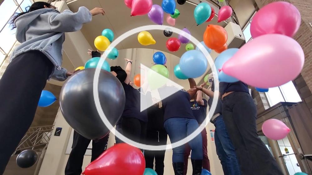 Students standing among falling balloons with a play symbol superimposed over the image