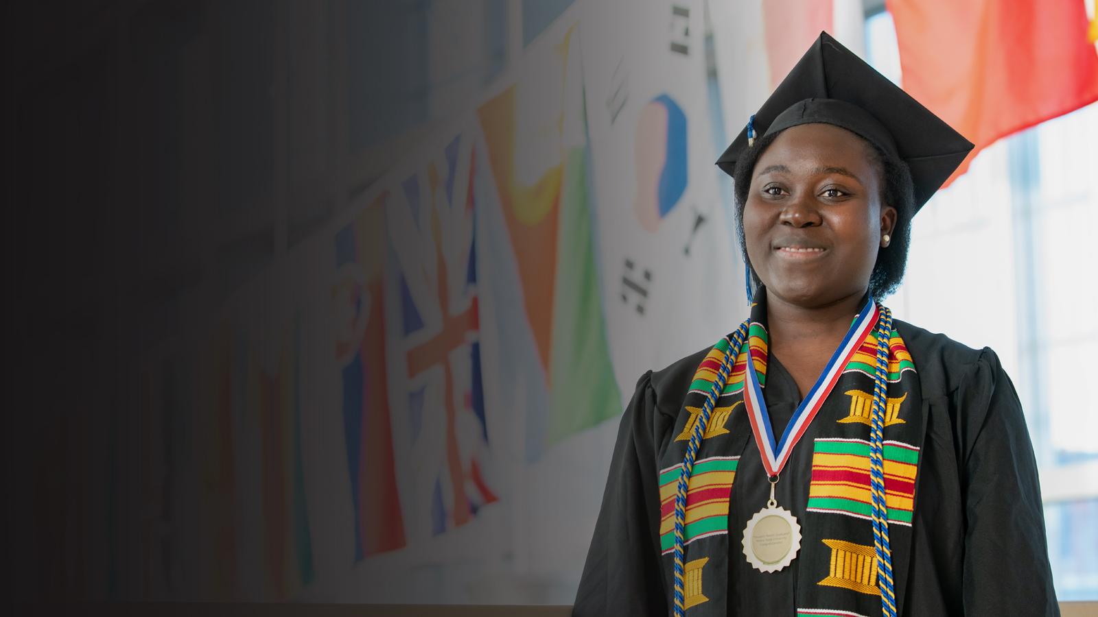 A person in cap and gown stands in front of the flags hanging in New Main