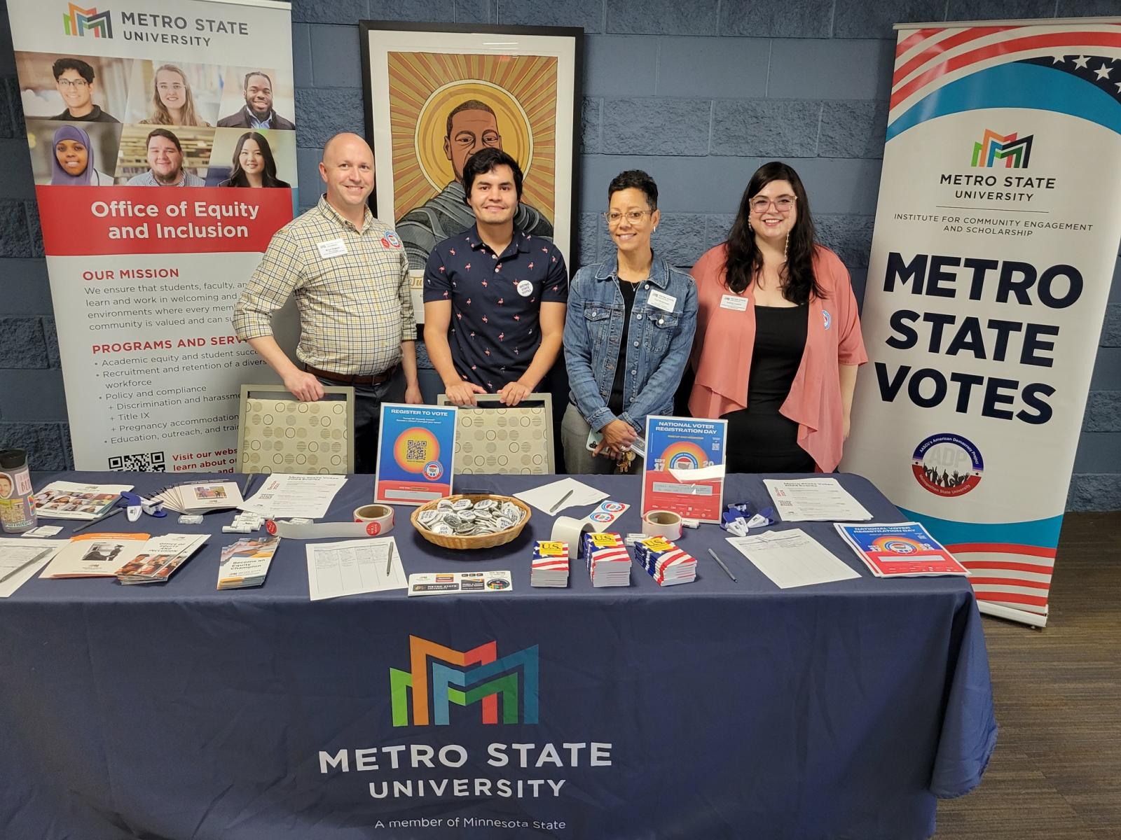 A group of people stand behind a table with buttons and flyers next to a Metro State Votes banner