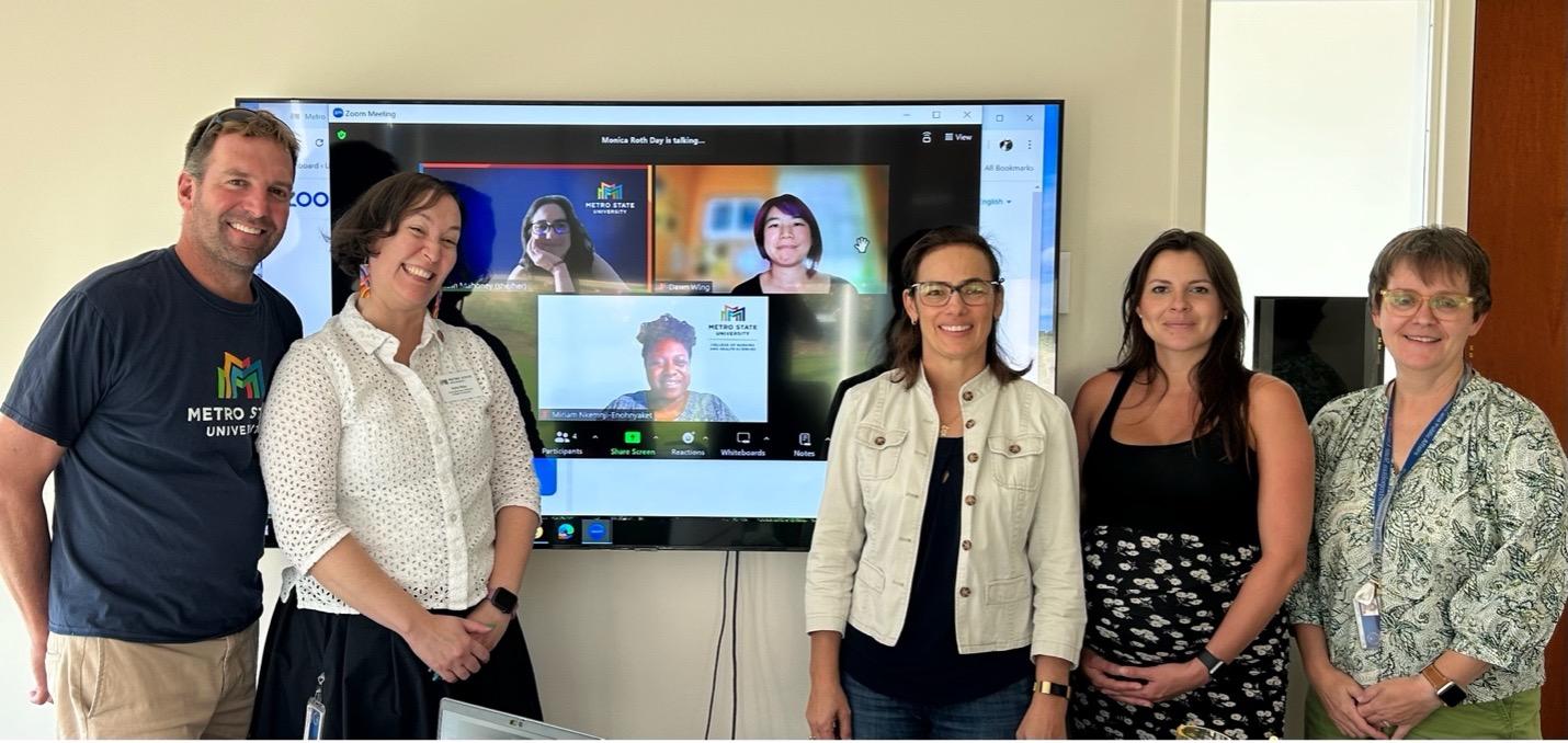 A group of people pose near a screen where three other people look on through video conference screens